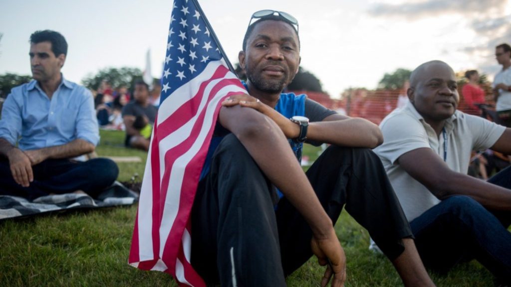 Man in crowd sitting on lawn with American flag resting on his shoulder (© Brianna Soukup/Portland Press Herald/Getty Images)